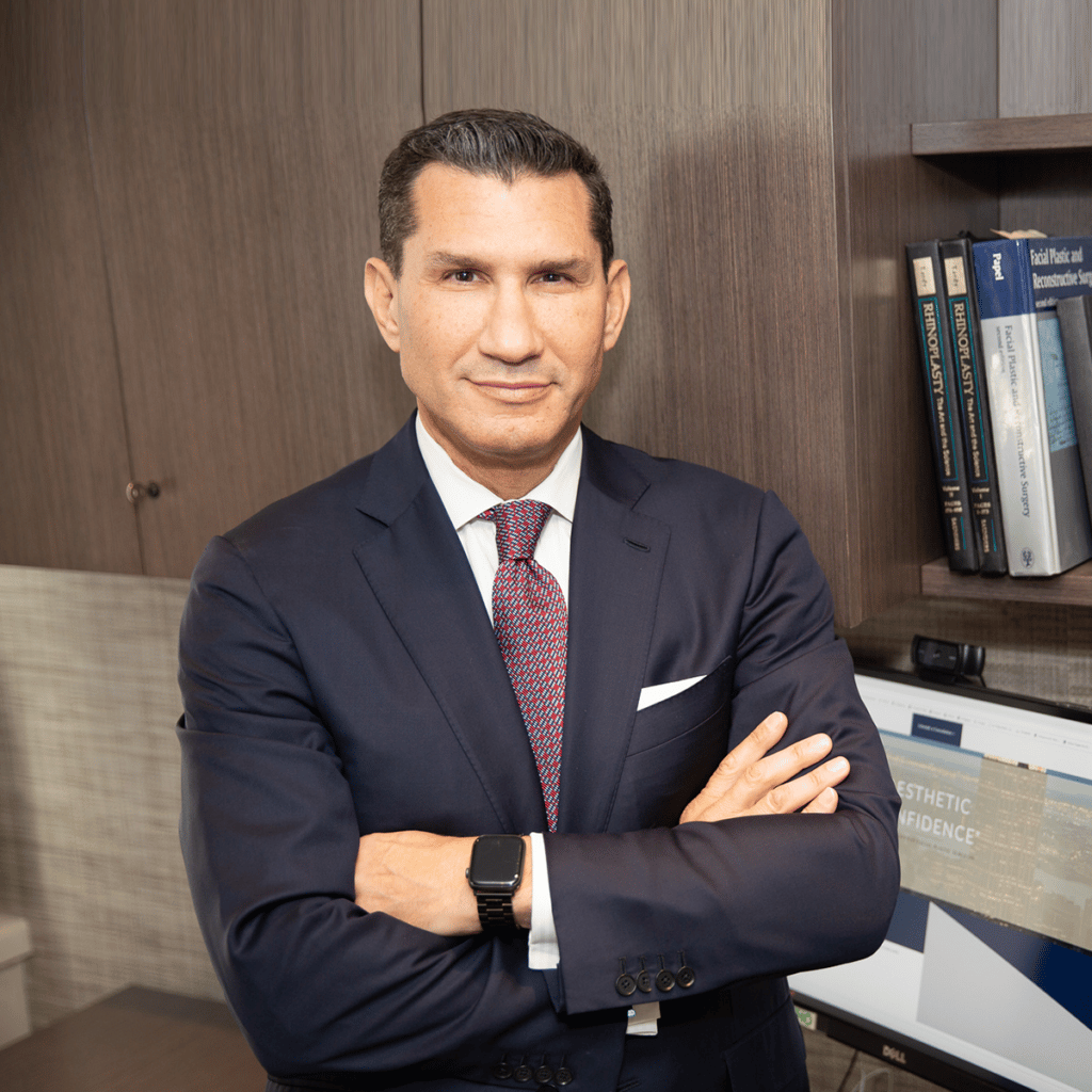 Professional portrait of Dr. Miller in a navy suit with a red tie and white pocket square, arms crossed, standing in an office with wood shelves and a computer monitor behind him.