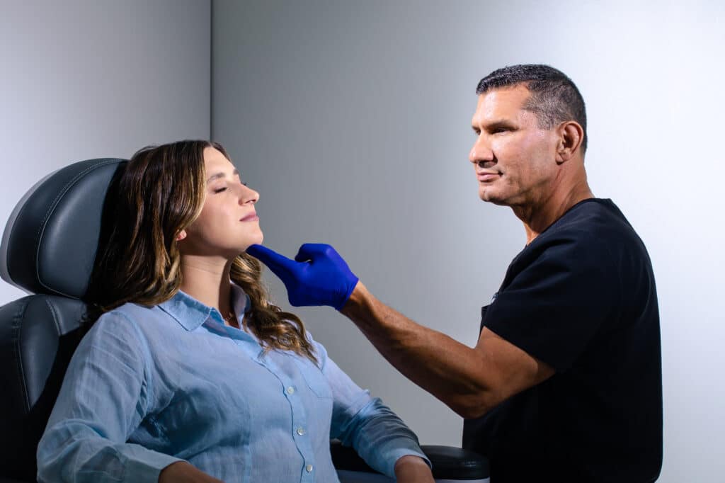 Dr. Miller in a dark shirt wearing a blue glove examining a seated woman’s face in a clinical setting.